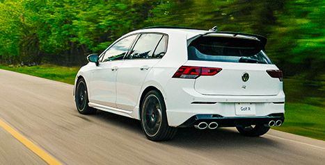 Dynamic rear view of a white Volkswagen Golf R driving on a country road, showing the roof spoiler and quad exhaust.