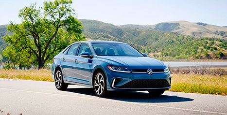 A blue sedan is driving on a road near a lake, with green rolling hills in the background.