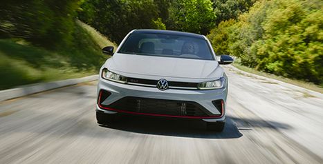 Front view of a white Volkswagen Jetta GLI sedan driving quickly on a winding road surrounded by trees.