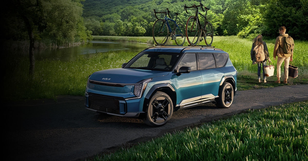 A blue Kia EV9 electric SUV with two bicycles on a roof rack, parked on a path next to a river in a lush, green forest.