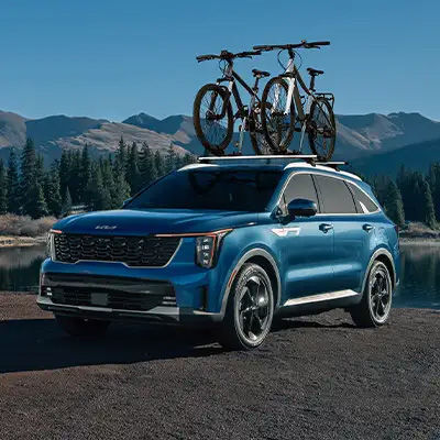A blue Kia Sorento SUV with two bicycles on a roof rack, parked on a gravel road next to a lake with mountains in the background.
