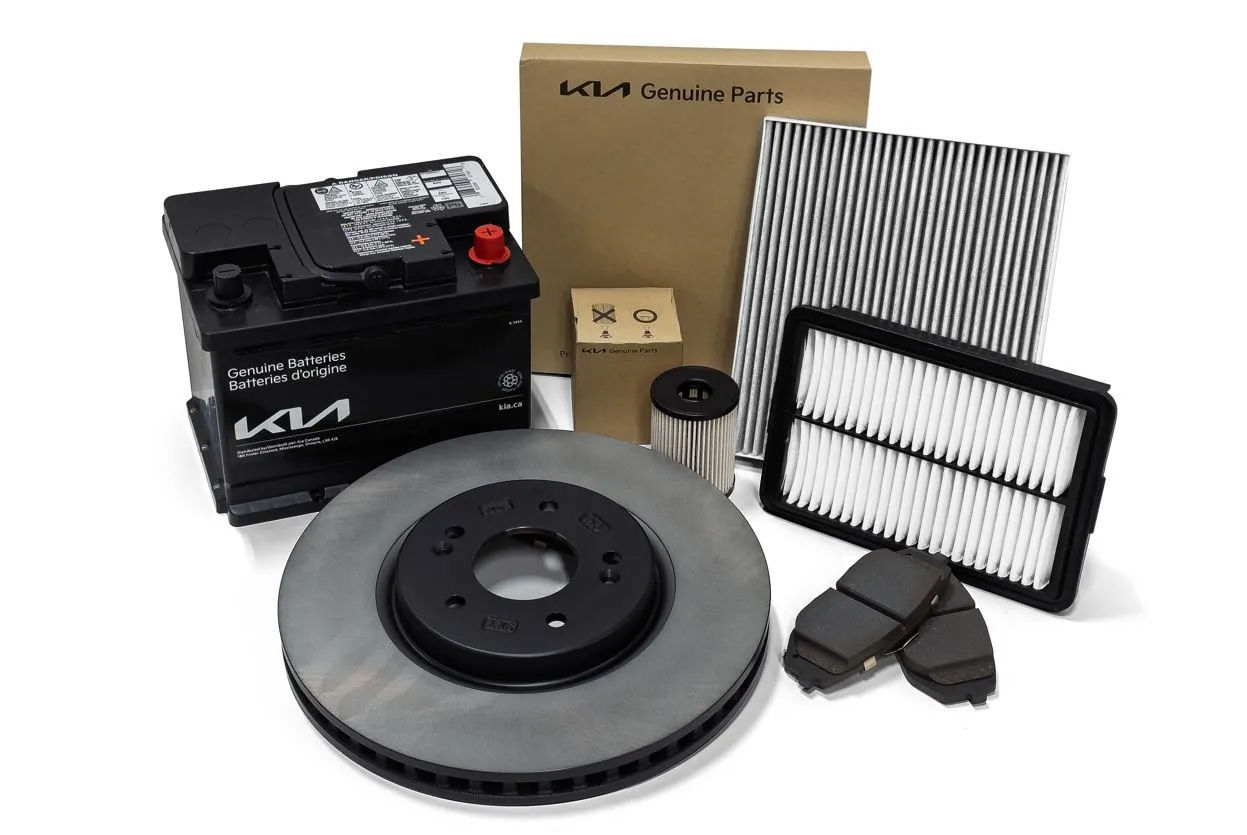 A collection of Kia Genuine Parts is displayed on a white background. The parts include a car battery, a brake disc, brake pads, an engine air filter, a cabin air filter, and an oil filter in its box. A larger Kia Genuine Parts box is also visible in the background.