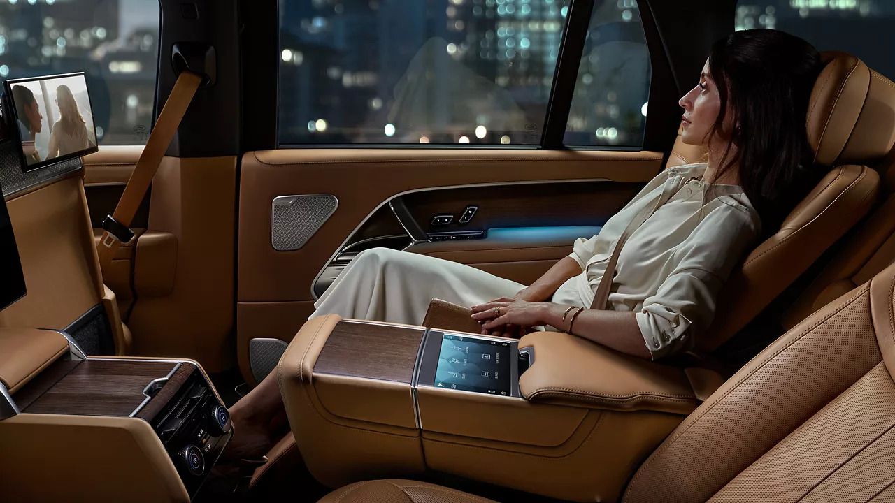Relaxed woman in the rear of a luxury tan leather cabin, with a rear-seat entertainment screen.