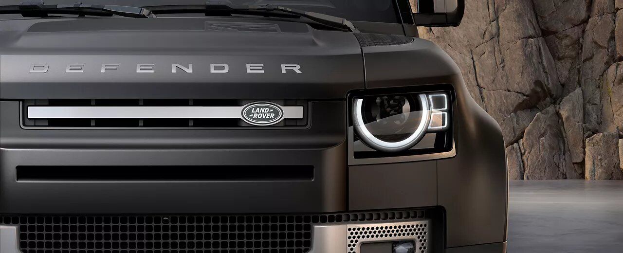 Close-up of a matte black Land Rover Defender front grille, featuring the Land Rover badge, "DEFENDER" lettering on the hood, and the signature circular LED headlight.
