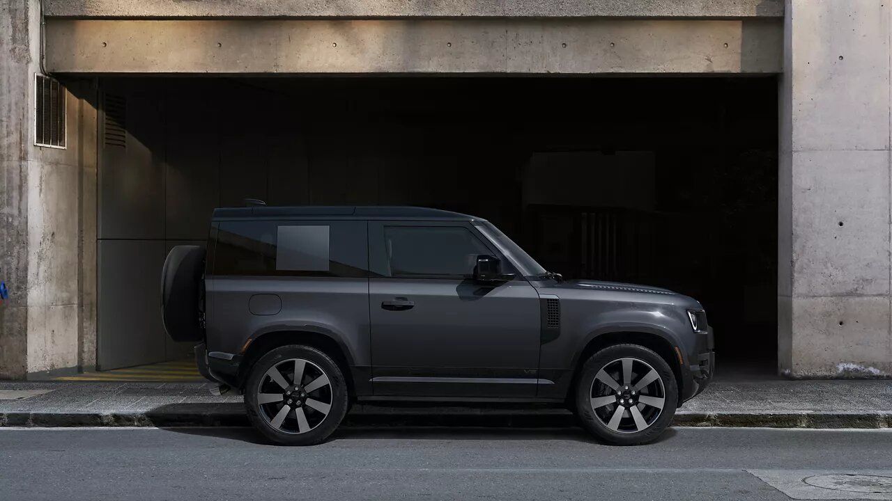 Side profile of a charcoal grey Land Rover Defender 90 (3-door short wheelbase model) parked in front of a modern urban concrete building.
