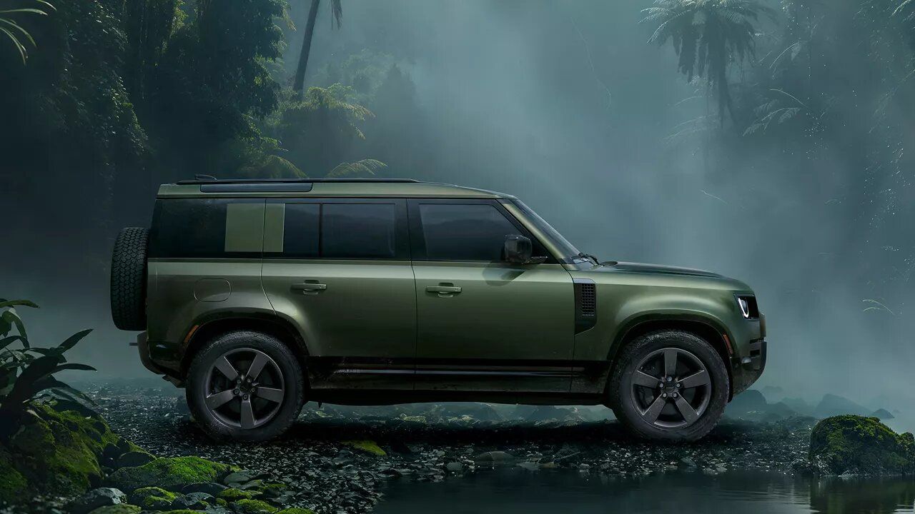 An olive green Land Rover Defender parked in a dense, misty tropical jungle surrounded by ferns and palm trees.