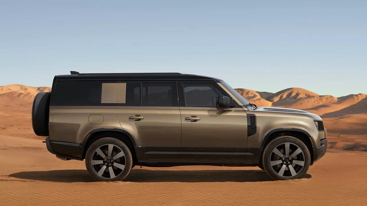 Side view of a bronze/gold Land Rover Defender in a sand dune desert under bright sunlight.