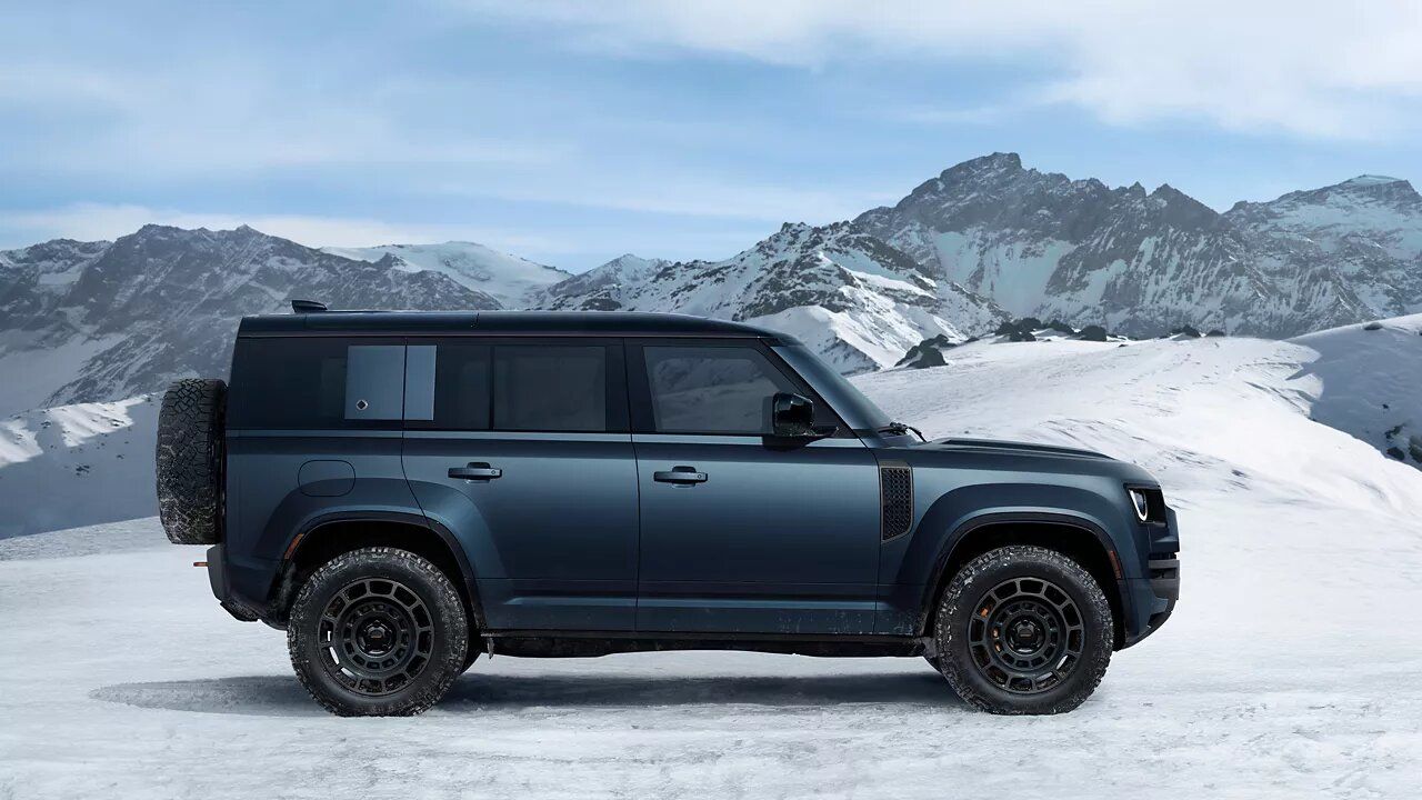 Side profile view of a dark blue Land Rover Defender parked in a snowy mountain landscape under a clear sky.