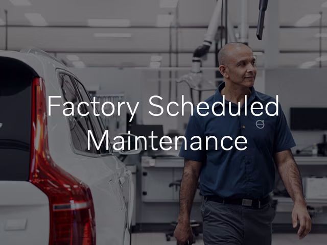 A male technician in a blue Volvo uniform walks through a bright, clean service bay, with the rear of a white Volvo XC90 on the left. The text "Factory Scheduled Maintenance" is overlaid on the image.