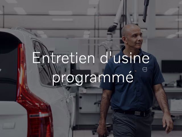 Un technicien dans un uniforme bleu Volvo marche dans un centre de service propre et lumineux, avec l'arrière d'un VUS Volvo XC90 blanc à gauche. Le texte « Entretien d'usine programmé » est superposé sur l'image.