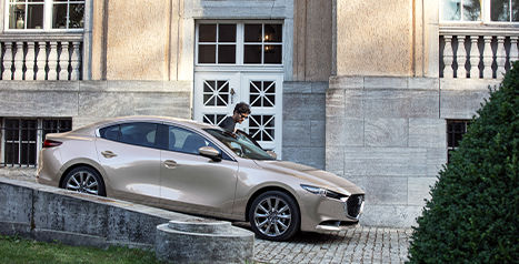 A person is getting into the driver's side of a gold-colored Mazda 3 sedan parked on a cobblestone surface in front of a grand building.