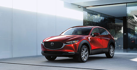 A shiny red Mazda CX-30 SUV parked on a paved driveway in front of a modern building with white walls and large windows.