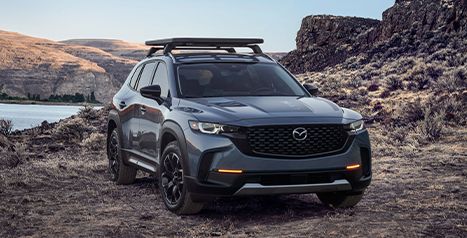  A dark grey Mazda CX-50 SUV featuring a roof rack, parked on a dirt road in a rugged, rocky landscape next to a body of water.