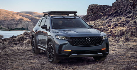 A dark grey Mazda CX-50 SUV featuring a roof rack, parked on a dirt road in a rugged, rocky landscape next to a body of water.