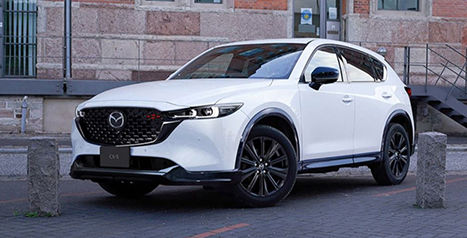 A white Mazda CX-5 SUV parked on a city street with a large, classic stone building in the background.