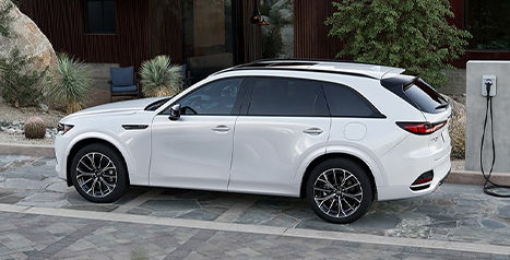 A white Mazda CX-70 SUV, shown in profile while parked in a paved driveway and plugged into an electric vehicle charging station.