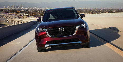 A front view of a deep red Mazda CX-90 SUV driving along a multi-lane highway, with a city skyline visible in the background.