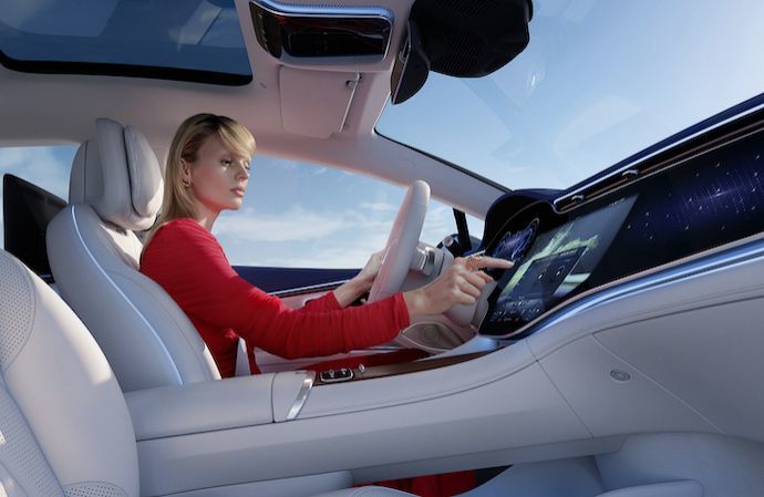 A woman in a red dress sitting at the steering wheel uses the large dashboard touchscreen display inside a modern car.