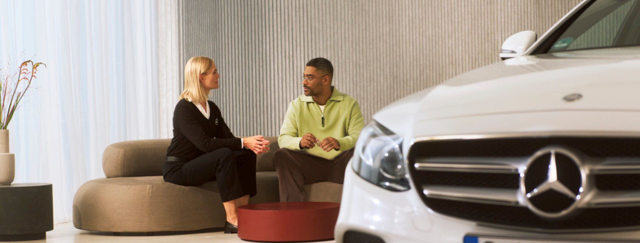 A service advisor and a customer talking on a sofa next to a white Mercedes-Benz.