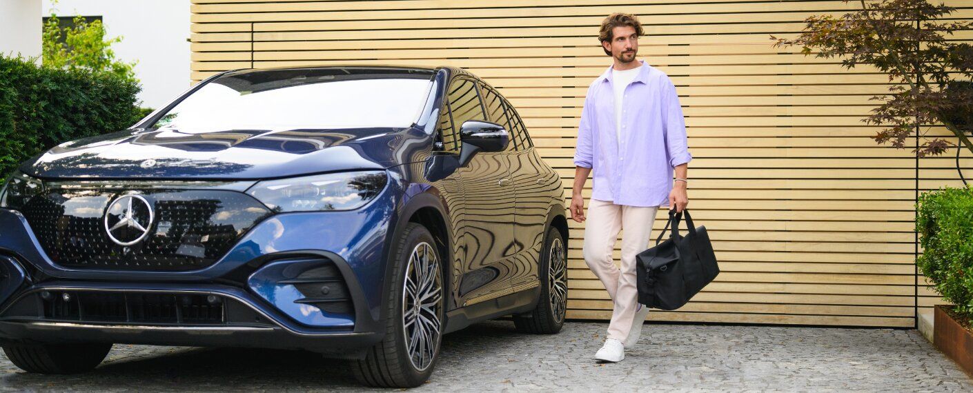 A man holding a duffel bag walking next to a dark blue Mercedes-Benz SUV.