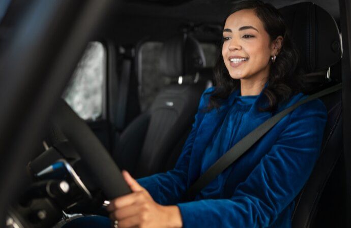 A smiling woman in a blue velvet blazer driving a car with her hands on the steering wheel.