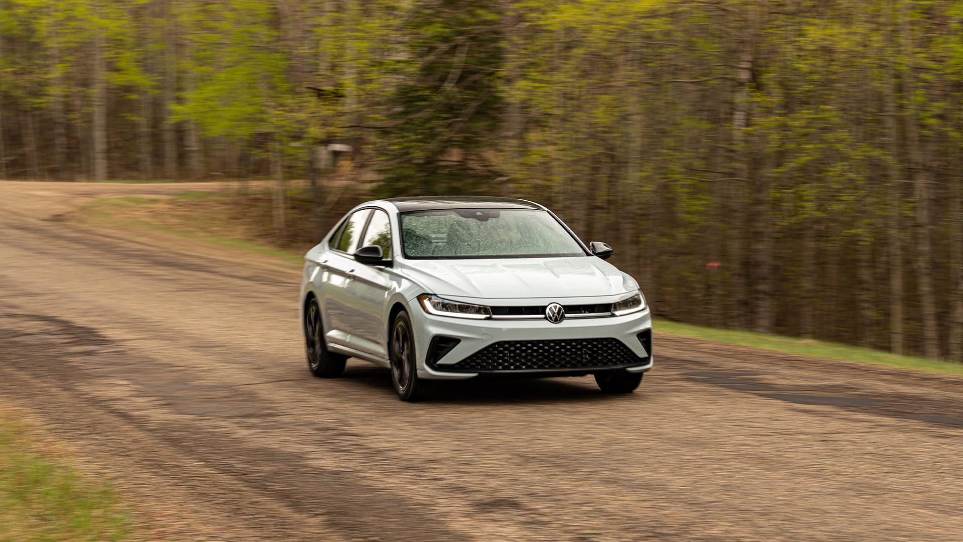 2027 Volkswagen Jetta driving on a dirt road in Reflex Silver Metallic front three-quarter view with forest backdrop