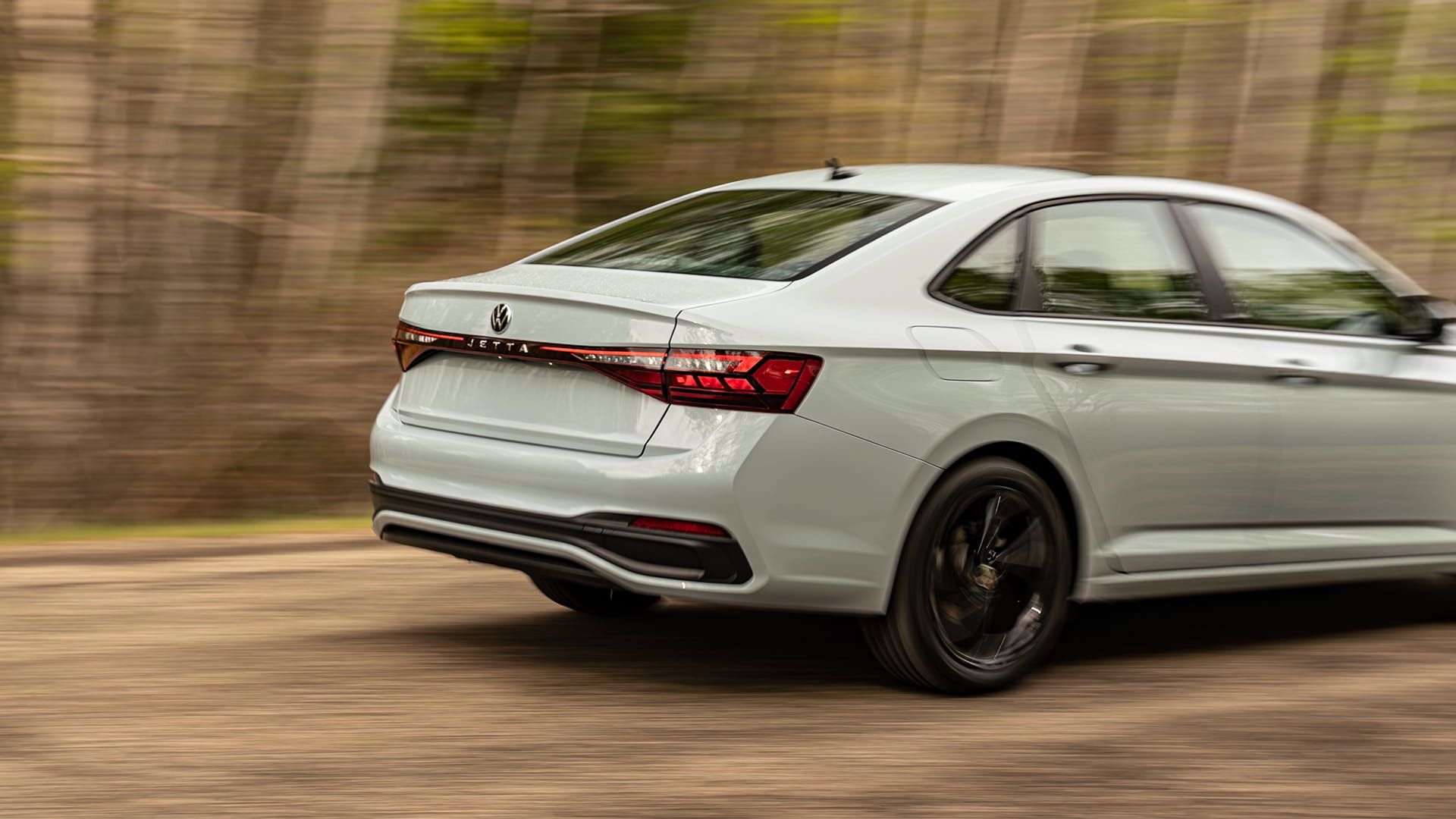 2027 Volkswagen Jetta rear view in motion on a tree-lined road with integrated spoiler and LED tail lights