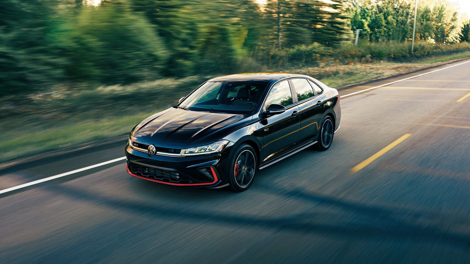 Black Jetta GLI driving on an open road with autumn trees in the background.