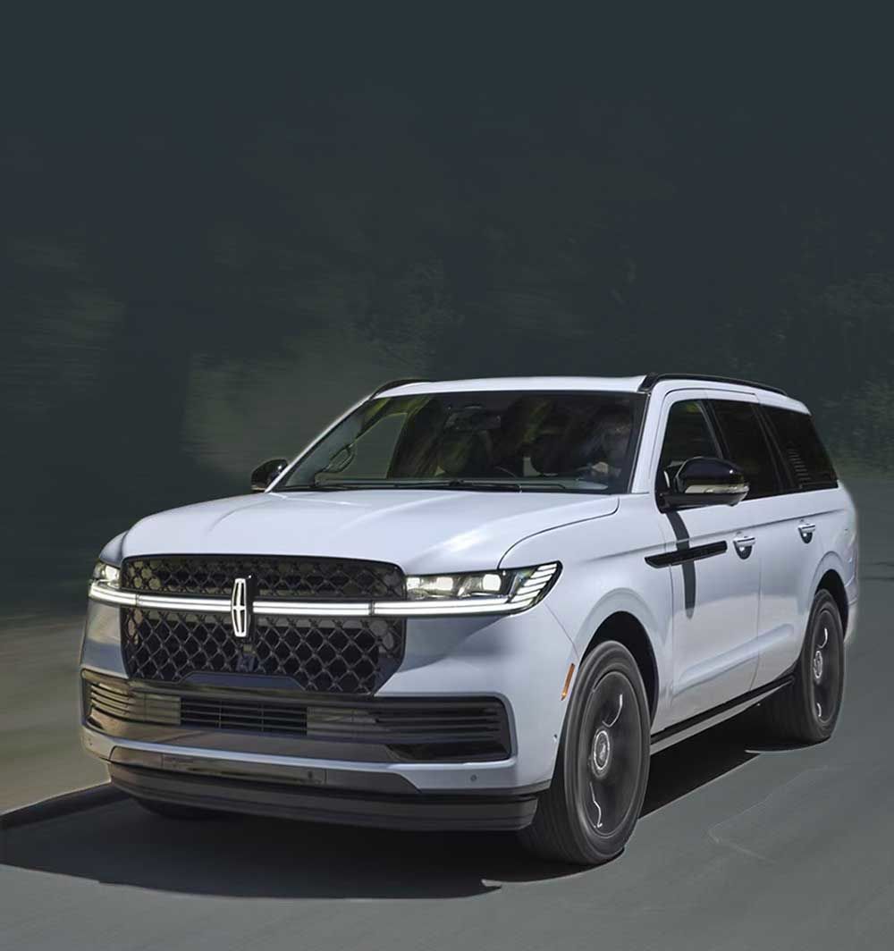 A light gray Lincoln Navigator SUV drives on a tree-lined road. The car is seen from the front, with motion blur suggesting speed.