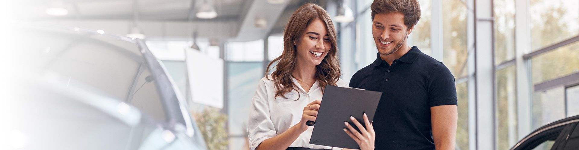 A smiling female salesperson and a male customer look at a clipboard together inside a bright car dealership showroom.