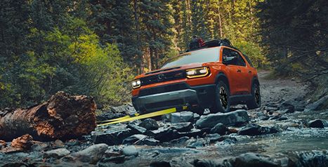 Un Honda Passport orange traversant un ruisseau rocheux dans un décor forestier, avec une sangle de remorquage jaune attachée au crochet avant.