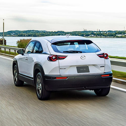 View of MX-30 EV’s rear as it drives down a riverside road. 