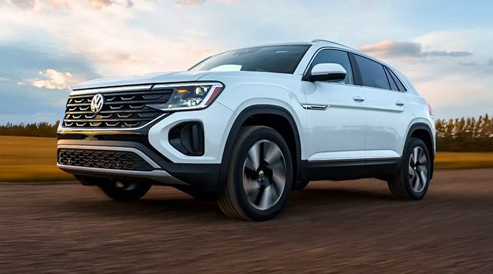 White Volkswagen Atlas Cross Sport in motion, front three-quarter view on a dirt road under a sunset sky.