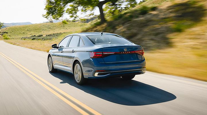 Rear view of the blue Jetta driving on a scenic road with a large tree and grassy hills in the background.