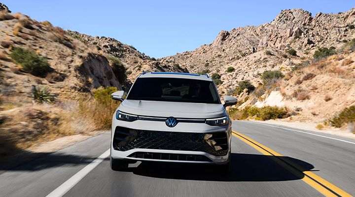 Front view of a light grey Volkswagen Tiguan or SUV driving on a road in a desert environment under a clear blue sky.