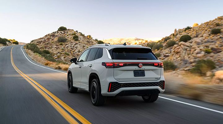 Rear view of a light grey Volkswagen Tiguan in motion on a desert road, showing the taillights and the "Tiguan" badge on the liftgate.