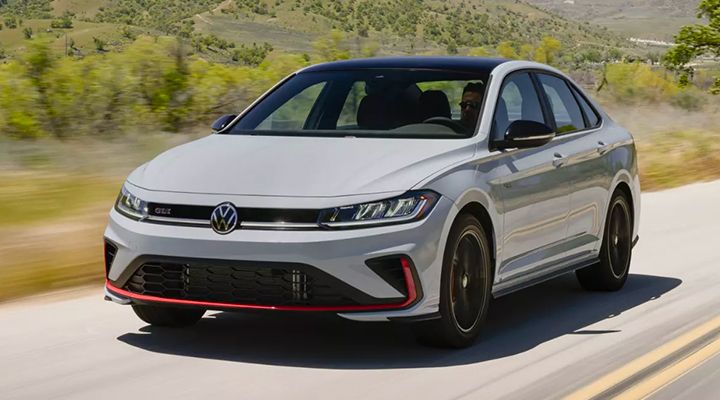 A white Volkswagen Jetta GLI  drives along a scenic road, with lush green hills in the background.