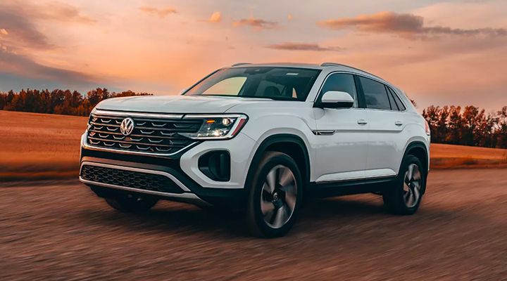 White Volkswagen Atlas Cross Sport in motion, front three-quarter view on a dirt road, under an orange and pink sunset sky.