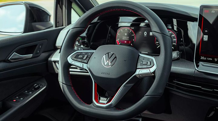 Close-up on the Golf GTI's sport steering wheel with red stitching and the GTI badge, with the digital dashboard visible in the background.