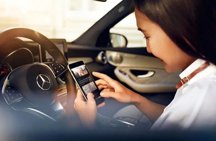 A woman sitting at the steering wheel of a car looking at an app on her smartphone.