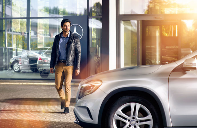 A man in a leather jacket walking outside a Mercedes-Benz dealership next to a display car.