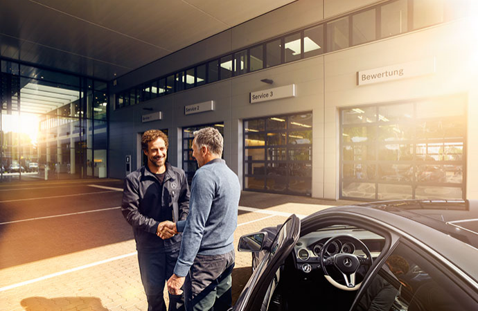A mechanic shaking hands with a customer in front of a car service center workshop bays.