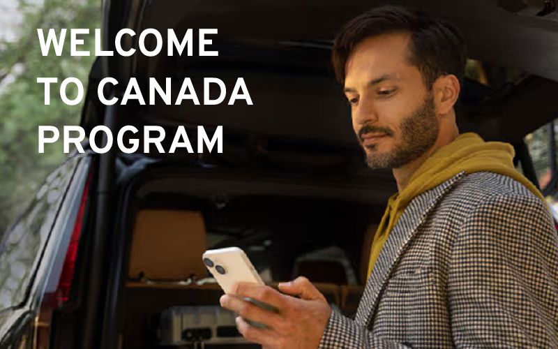 Man standing near the open trunk of a vehicle looking at his smartphone, with the text "WELCOME TO CANADA PROGRAM" overlayed.