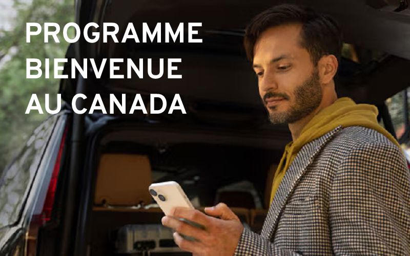 Homme près du coffre ouvert d'un véhicule, regardant son téléphone intelligent, avec le texte "PROGRAMME BIENVENUE AU CANADA" superposé.