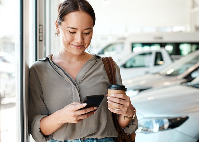 Une femme souriante se tient dans un concessionnaire automobile, regardant son téléphone intelligent tout en tenant un gobelet de café.