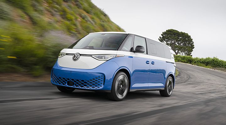 Une fourgonnette électrique Volkswagen ID. Buzz bicolore bleu et blanc roule sur une route sinueuse bordée de verdure.