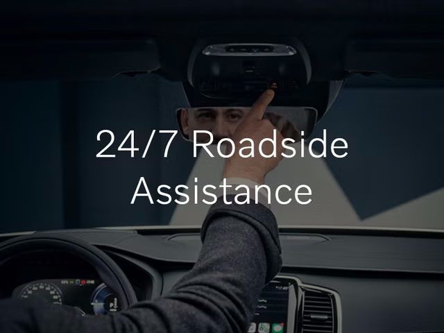 From the driver's seat, a person reaches up to press the "SOS" button on the overhead console of a Volvo. The text "24/7 Roadside Assistance" is overlaid on the image.