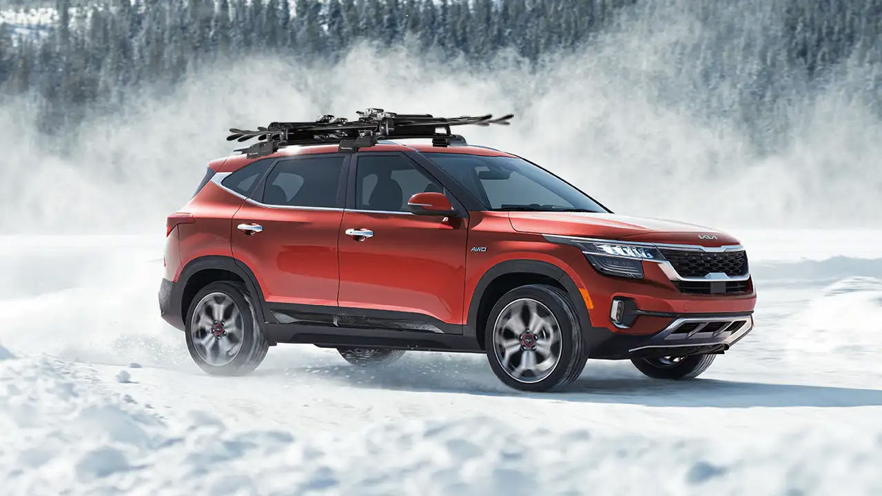 An orange Kia Seltos SUV with skis on its roof rack, driving through a snowy landscape and kicking up snow.