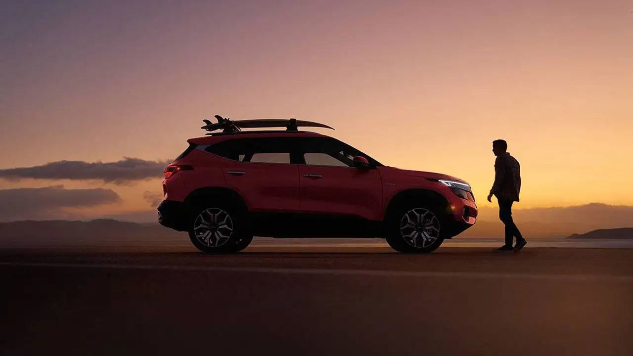  A red Kia Seltos SUV with a surfboard on its roof rack, parked on a beach at sunset as a person walks toward it.