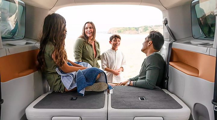 Une famille est assise dans le grand espace de chargement arrière ouvert d'un fourgon sur une plage, en train de converser.